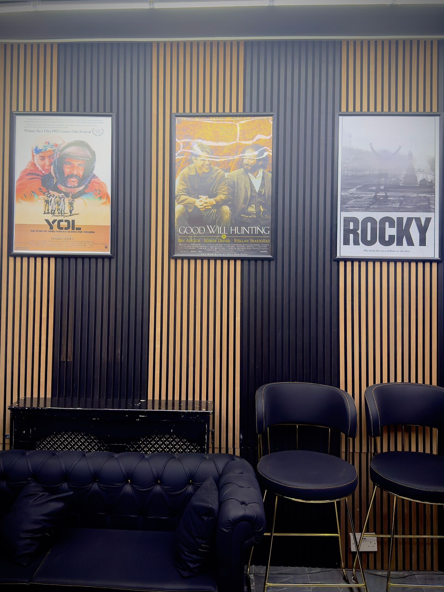Waiting lounge at Xalo Barbers with slatted wood wall, framed film posters, and navy leather seating